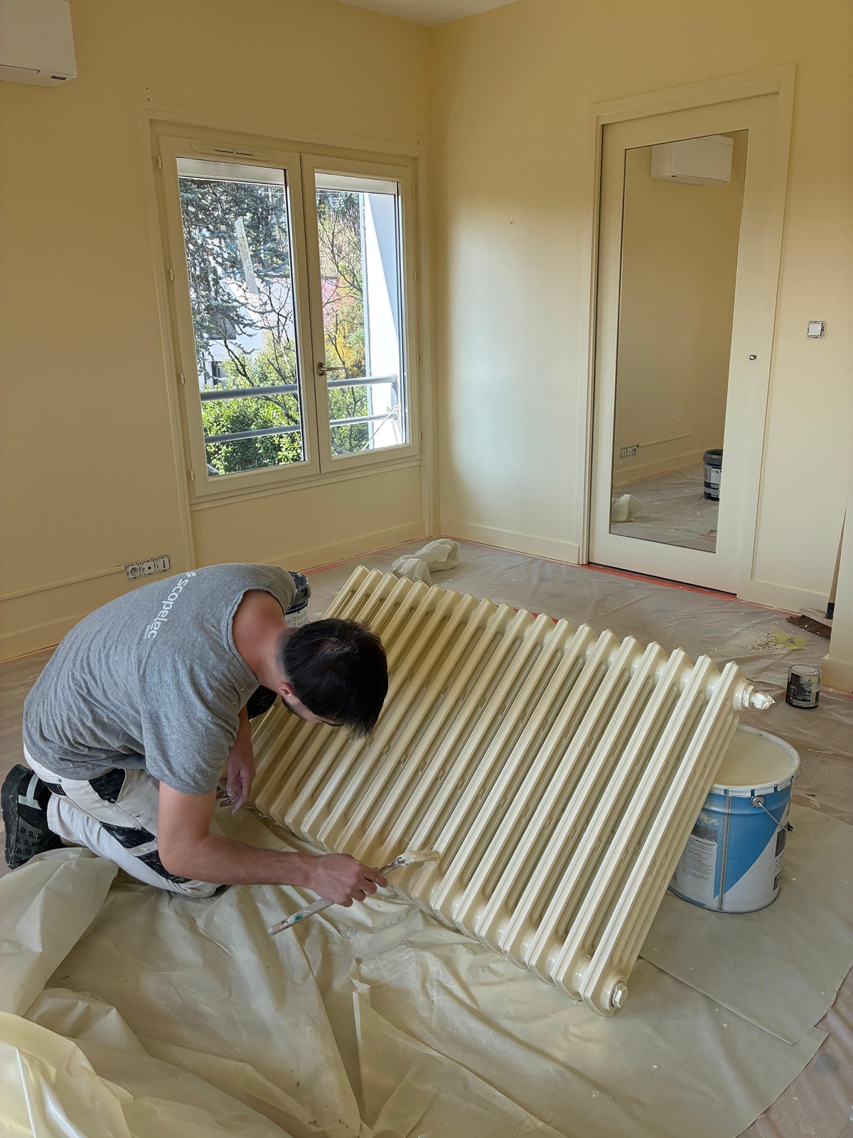 Photo d'un ouvrier en train de peindre un radiateur en fonte sur un sol protégé par une bâche en plastique.
